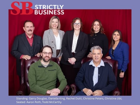 Group portrait labeled ‘Strictly Business’ showing seven professionals against a red backdrop. Standing (left to right): Garry Douglas, Danielle King, Rachel Dutil, Christine Peters, Christina Ubl. Seated: Aaron Roth and Todd McCarthy.