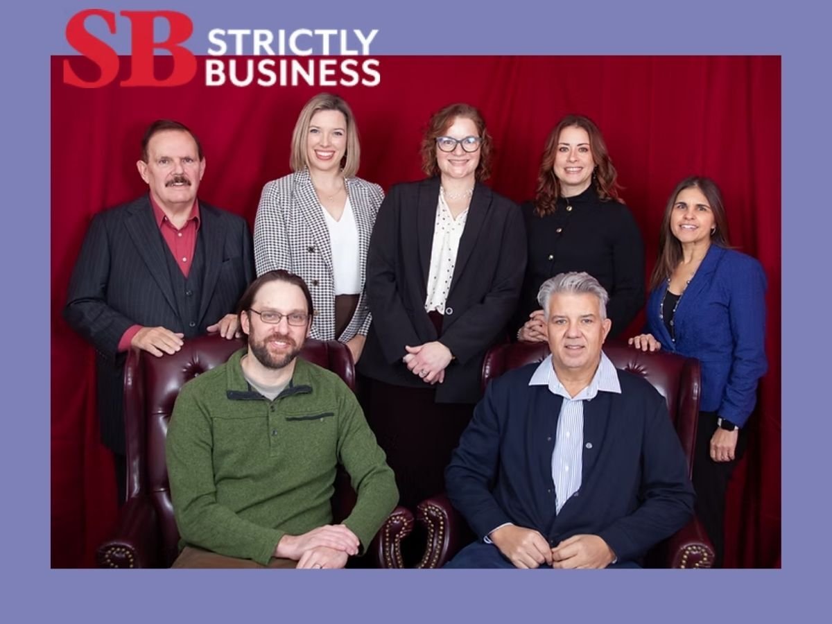 Group portrait labeled ‘Strictly Business’ showing seven professionals against a red backdrop. Standing (left to right): Garry Douglas, Danielle King, Rachel Dutil, Christine Peters, Christina Ubl. Seated: Aaron Roth and Todd McCarthy.